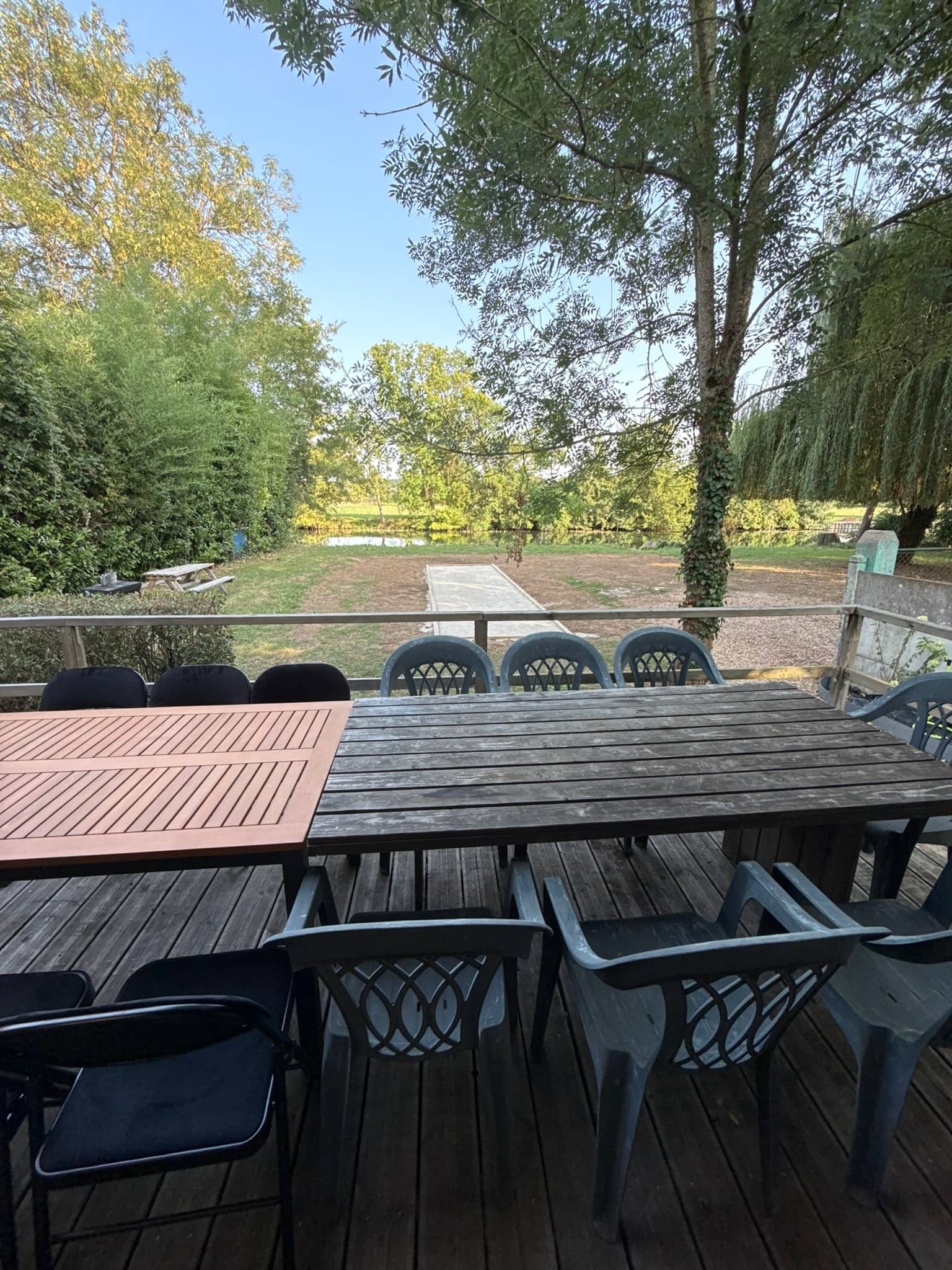 Terrasse couverte du chalet : grande table en bois, puis la pelouse, le terrain de pétanque et le Loir au fond du jardin